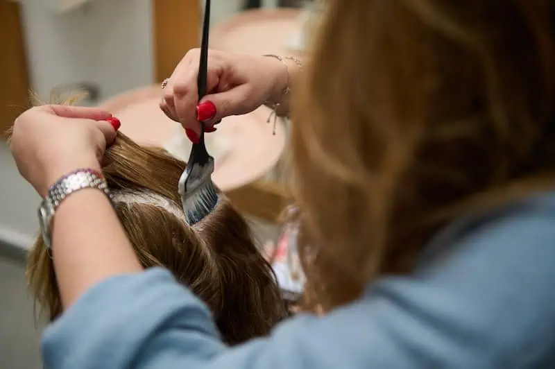 6 napak, ki jih delamo pri uporabi lasnih podaljškov in kako jih preprečiti 7 V salonu oseba v modri srajci natančno nanaša barvo za lase s čopičem na korenine rjavih las. Njihov rdeč lak za nohte doda piko na i, saj mojstrsko delujejo in zagotavljajo, da je vsak pramen deležen pozornosti.